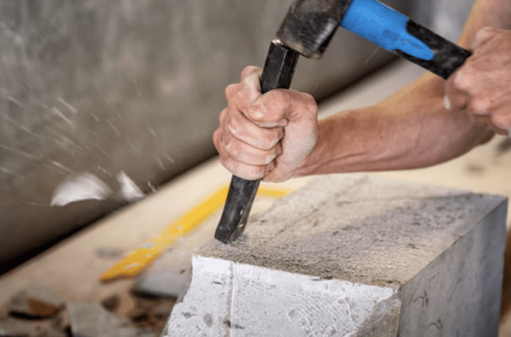 Close-up of hands chiseling a natural stone block with a hammer and chisel. The person, in a sleeveless shirt, embodies the timeless beauty of craftsmanship as stone chips fly off. The blurred background draws focus to this architectural creation in progress.